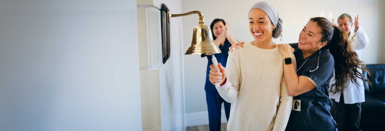Female cancer patient wearing head scarf smiles while ringing bell and being cheered on by Asian female nurse and other healthcare workers in background